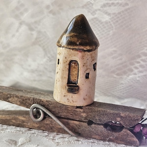 May include: A small, ceramic tower with a brown, cone-shaped roof and a beige body. The tower has a rectangular door and small square windows. It rests on a wooden clothespin, with a silver wire spring.