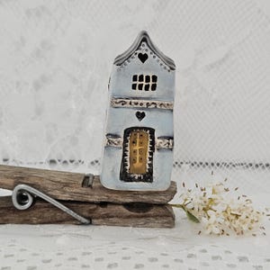 May include: A miniature ceramic house painted in light blue with a black roof and heart details. The house has a small door and window. It is mounted on a wooden clothespin, with a sprig of white flowers in the foreground.