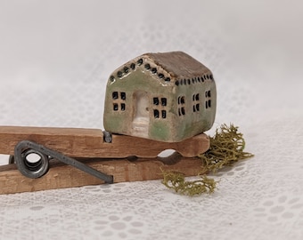 May include: A small green and brown ceramic house with black windows and a white door, sitting on a wooden clothespin.