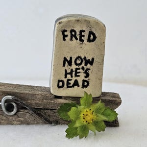 May include: A small, off-white, tombstone-shaped ceramic piece with the black text "FRED NOW HE'S DEAD" printed on the front. It is attached to a weathered, dark brown wooden clothespin, and a small green leaf with a yellow center sits in front.