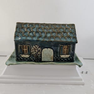 May include: A ceramic butter dish shaped like a small house with a dark blue, textured roof and walls. The front features a central arched door, two windows with blue shutters, and a small tree. The house sits on a matching rectangular base.