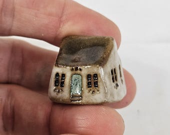 May include: A small, white ceramic house with a brown roof and black windows. The house has a green door and a small porch.