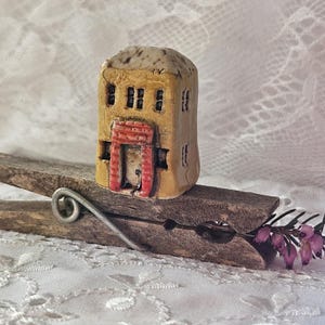 May include: A miniature ceramic house with a tan exterior, small windows, and a red door frame. The house is perched on a wooden clothespin. The roof has a textured, speckled appearance. A sprig of purple flowers is visible.