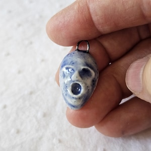 May include: A small, blue and white ceramic pendant in the shape of a screaming face. The pendant has a dark blue bail and a speckled pattern. The face has open eyes and a wide open mouth.