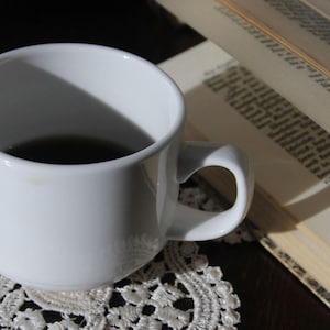 Op de afbeelding: Een witte keramische mok gevuld met een donkere vloeistof, mogelijk koffie of thee, staat op een wit kanten kleedje. Een open boek met zichtbare tekst is gedeeltelijk op de achtergrond te zien.