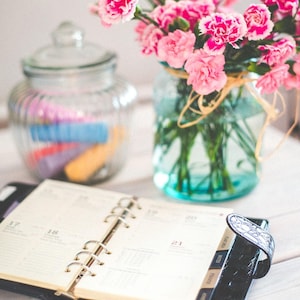 May include: An open planner with a black cover sits on a white surface, next to a glass jar filled with colorful items and a vase of pink flowers. The flowers are in a blue-tinted glass jar tied with twine.