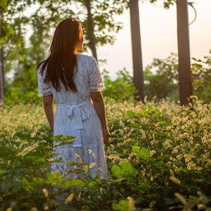 May include: A white eyelet dress with short sleeves and a tie-back waist. The dress is knee-length and the person is standing in a field of tall grass. The background includes trees and sunlight.