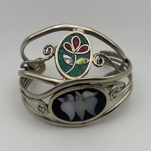 May include: Silver-toned cuff bracelet with two oval inlays. One inlay features a green mosaic with a red and pink flower design. The other inlay has a black background with three white, iridescent petal shapes. The bracelet has a decorative, open design.