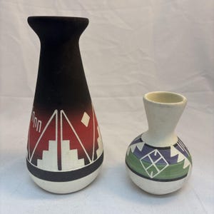 May include: Two ceramic vases. The taller vase features a black, red, and white color scheme with geometric step patterns. The smaller, rounder vase is cream-colored with green, blue, and white geometric designs, including a central diamond motif.