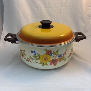 Puede incluir: Una olla de cocina redonda de metal con una base de color crema que presenta un patrón floral colorido de flores amarillas, naranjas y azules. La olla incluye una tapa de color amarillo brillante con un pomo marrón oscuro y dos asas laterales.