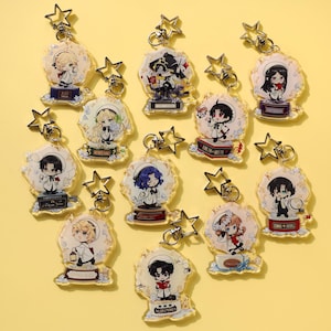 Könnte beinhalten: Eine Sammlung von Acryl-Schlüsselanhängern mit Anime-Charakteren. Jeder Schlüsselanhänger hat ein einzigartiges Design mit einem sternförmigen Verschluss. Die Charaktere stehen auf dekorativen Sockeln, einige mit Text.