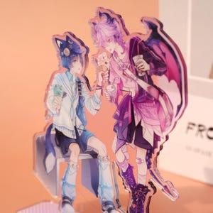 May include: Acrylic stand featuring two anime-style characters. One character has blue hair and a cat-ear hat, holding ice cream. The other character has purple hair and a dragon wing design, holding a drink. The artwork has a vibrant color palette.