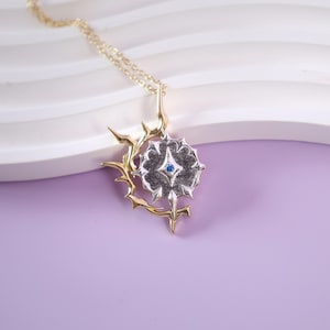 Könnte beinhalten: Halskette mit Anhänger aus Gold und Silber. Der Anhänger hat ein sternförmiges, dunkelgraues Zentrum mit einem kleinen blauen Edelstein, umgeben von einem goldenen, dornigen Rahmen. Die Halskette ist goldfarben.