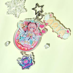 May include: A clear acrylic keychain featuring a colorful anime-style illustration of a character named Aria. The keychain includes a star-shaped clasp, a charm with the name "ARIA", and a smaller charm with the text "ANGEL OF COLLUSION".