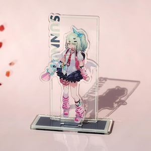 Könnte beinhalten: Ein klarer Acrylständer mit einem farbenfrohen Anime-Charakter. Die Figur hat grüne Haare, eine blaue Schleife und trägt ein weißes Hemd, eine rosa Jacke und einen schwarzen Rock. Das Wort "SUNNY" ist vertikal auf dem Ständer aufgedruckt.