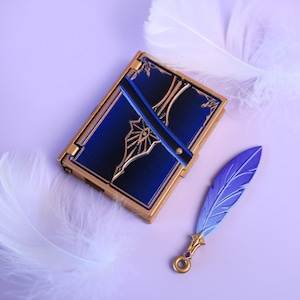 May include: A blue and gold decorative book with a matching blue and purple feather pen. The book has a gold border and a blue cover with gold filigree. The feather pen is blue and purple with a gold tip.