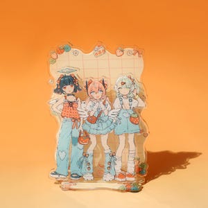 Könnte beinhalten: Ein klarer Acrylständer mit drei Anime-Charakteren mit Engelsflügeln. Die Charaktere tragen bunte Outfits mit Erdbeer- und Herzmotiven. Der Hintergrund ist hellorange.