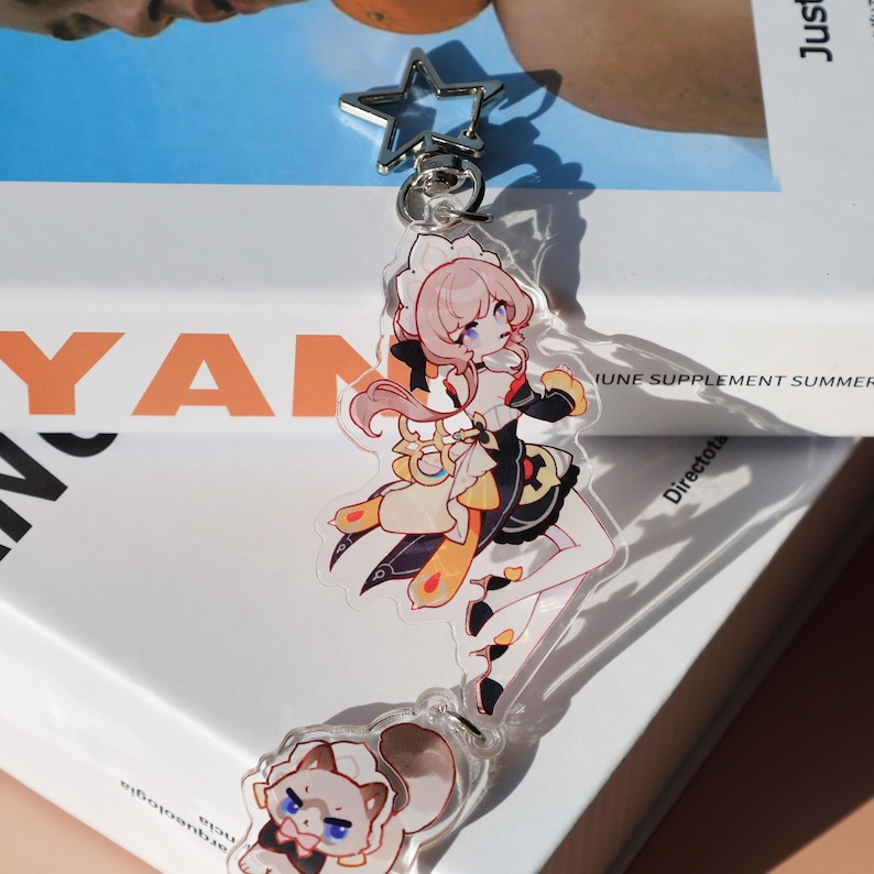 May include: A clear acrylic keychain featuring a colorful anime-style character with pink hair, wearing a black and yellow outfit. The keychain has a star-shaped clasp and a second charm with a cartoon animal.
