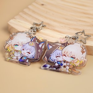 May include: Two clear acrylic keychains featuring anime-style characters in a hugging pose. One keychain has a star-shaped clasp, the other has a heart-shaped clasp. The characters have light hair, and are wearing detailed outfits. The keychains are on a light brown wooden surface.