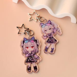 Könnte beinhalten: Zwei Anime-Stil-Schlüsselanhänger aus Acryl. Jeder zeigt ein Mädchen mit lila Haaren, das ein schwarzes und lila Outfit und eine Sonnenbrille trägt. Die Schlüsselanhänger haben silberne sternförmige Verschlüsse. Der Hintergrund ist hellpfirsichfarben.