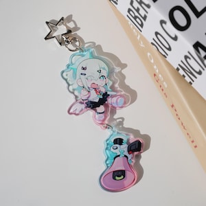 Op de afbeelding: Een heldere acryl sleutelhanger met een cartoonkarakter met lichtblauw haar en een zwarte rok. De sleutelhanger heeft een stervormige sluiting en een tweede bedel met een megafoondesign. De kleuren zijn lichtblauw, roze en zwart.