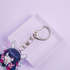 May include: A clear acrylic keychain featuring a cartoon character with dark blue hair, pink accents, and white wings. The keychain has a silver-colored clasp and chain. The background is a light purple.