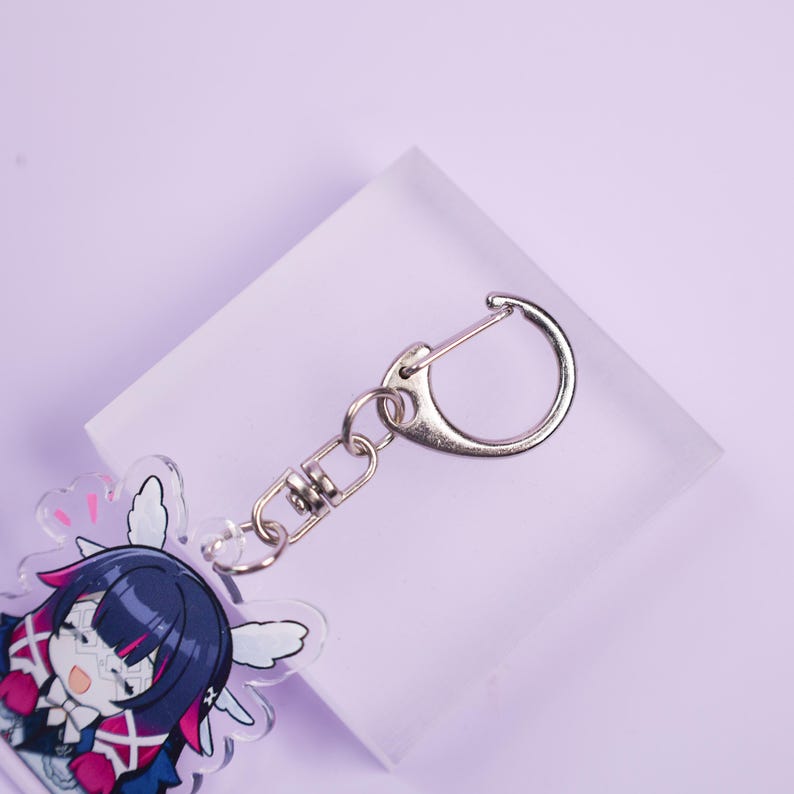 May include: A clear acrylic keychain featuring a cartoon character with dark blue hair, pink accents, and white wings. The keychain has a silver-colored clasp and chain. The background is a light purple.