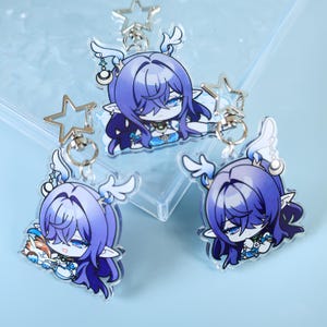 Könnte beinhalten: Drei Acryl-Schlüsselanhänger mit einem blauhaarigen Anime-Charakter mit elfenartigen Ohren und einem sternförmigen Schlüsselanhänger. Der Charakter trägt ein blaues Outfit mit weißen Akzenten. Die Schlüsselanhänger liegen auf einer hellblauen Oberfläche.