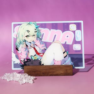 Può includere: Un espositore in acrilico in stile anime colorato con un personaggio con i capelli verde chiaro e una giacca a righe rosa e bianche. L'espositore è sostenuto da una base in legno e presenta la scritta "Sanna!" in lettere rosa. Una collezione di cristalli trasparenti è di fronte.