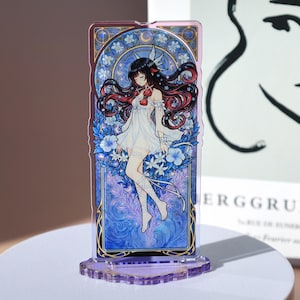 Può includere: Supporto decorativo in acrilico con un'illustrazione dettagliata di una donna in abito bianco con lunghi capelli neri e riflessi rossi. L'opera è ambientata su uno sfondo floreale e celeste blu, con una base viola.