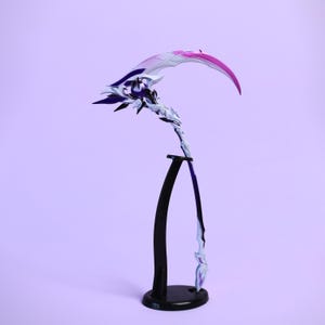 Könnte beinhalten: Eine dekorative Sense mit gebogener Klinge, die einen Farbverlauf aus Weiß, Lila und Rosa aufweist. Der Griff ist weiß mit lila Akzenten und floralen Details. Sie wird auf einem schwarzen Ständer vor einem lavendelfarbenen Hintergrund ausgestellt.