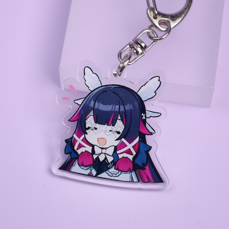 May include: A clear acrylic keychain featuring a cartoon character with dark blue hair, pink accents, and white wings. The character has a surprised expression. The keychain includes a silver-colored clasp.