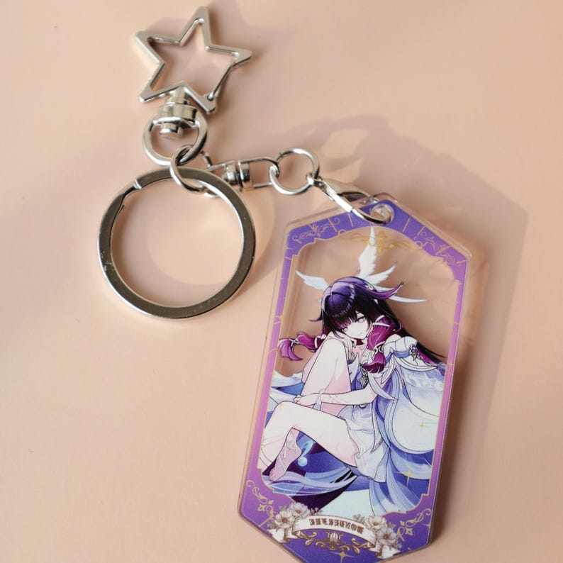 May include: A silver-colored keychain with a star-shaped clasp and a clear acrylic charm. The charm features a colorful anime-style illustration of a character in a purple and white outfit. The keychain is attached to a silver key ring.