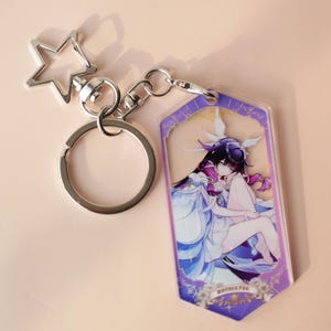 May include: A silver-colored keychain with a star-shaped charm and a clear acrylic tag. The tag features a colorful anime-style illustration of a character in a white and blue dress with purple accents. The keychain is designed for attaching to keys or bags.