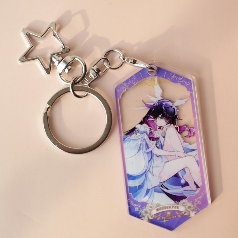 May include: A silver-colored keychain with a star-shaped charm and a clear acrylic tag. The tag features a colorful anime-style illustration of a character in a white and blue dress with purple accents. The keychain is designed for attaching to keys or bags.