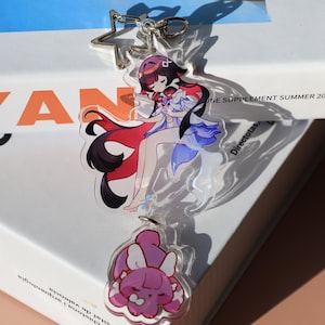 May include: A clear acrylic keychain featuring a colorful anime-style illustration of a character with red, black, and blue accents. The keychain includes a star-shaped clasp and a smaller charm with a pink creature design.