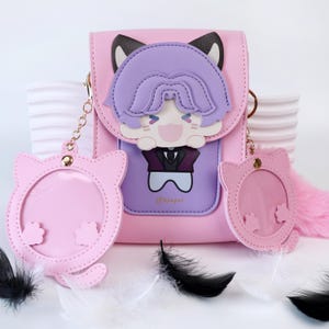 Könnte beinhalten: Rosa Umhängetasche mit einem Cartoon-Charakter-Design. Die Tasche hat ein Katzenohren-Design, einen lila Charakter mit einem lächelnden Gesicht und zwei rosa katzenförmige Schlüsselanhänger. Die Tasche hat eine kleine Tasche mit dem Text "@asaygrl".