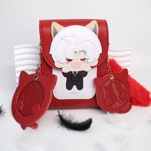 Könnte beinhalten: Rote Umhängetasche mit einem Cartoon-Charakter-Design. Die Tasche hat eine Klappe mit einer Charakterillustration, Fuchsohren-Akzenten und zwei passenden roten Schlüsselanhängern. Ein roter, flauschiger Schwanz und Federn sind ebenfalls enthalten.