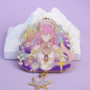 Puede incluir: Un pin de esmalte detallado que representa una figura femenina estilizada con cabello rosado y un tocado floral. El pin tiene un contorno dorado y está adornado con detalles morados, rosas y dorados. Un dije en forma de estrella cuelga de la parte inferior. El pin está sobre un fondo blanco texturizado.