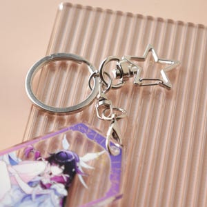 May include: Silver-toned keychain with a star-shaped clasp and a large ring. The keychain is attached to a rectangular charm with a purple border and a colorful anime-style illustration. The background is a light pink, ridged surface.