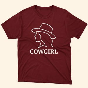 Può includere: T-shirt bordeaux con un contorno bianco della silhouette di una cowgirl con un cappello. La parola "COWGIRL" è stampata in bianco sotto. La maglietta ha maniche corte e girocollo.