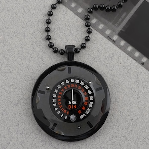 May include: A black pendant necklace featuring a circular pendant with a camera light meter design. The pendant has a black bezel with white and red markings, including ASA and DIN scales. The necklace chain is made of small, black beads.