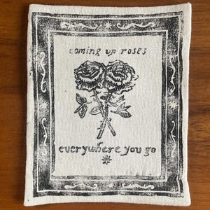 May include: Square, off-white fabric patch with a black floral design and text. The design features two roses and the phrases "coming up roses" and "everywhere you go" in a decorative frame.