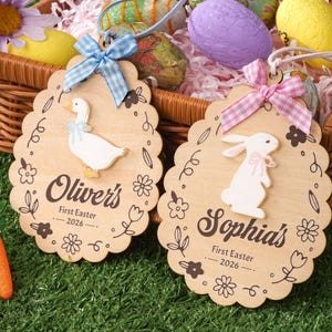 Puede incluir: Dos adornos de huevo de Pascua de madera con bordes festoneados. Uno presenta un pato blanco con un lazo azul, el otro un conejito blanco con un lazo rosa. Ambos tienen nombres y "First Easter 2026" impreso.