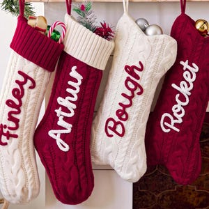 Puede incluir: Cuatro calcetines navideños de punto trenzado colgados en fila. Dos son de color crema con puños rojos y letras rojas, y dos son rojos con puños crema y letras blancas. Los nombres Finley, Arthur, Bough y Rocket están bordados en los calcetines.