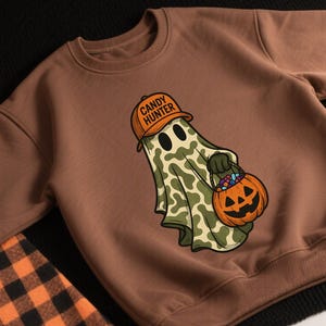 May include: Brown sweatshirt featuring a Halloween-themed graphic. The design includes a ghost wearing an orange hat with the text "CANDY HUNTER" and holding a pumpkin-shaped candy bucket. The ghost has a camouflage pattern. An orange pumpkin is visible in the upper left corner.