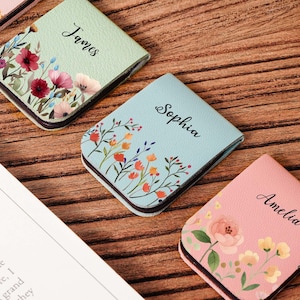 Może przedstawiać: Zbliżenie spersonalizowanych kwiatowych notatników w pastelowych kolorach. Notatniki są jasnozielone, jasnoniebieskie i różowe, każdy z imieniem w kursywie. Kwiatowe wzory obejmują czerwone, różowe i żółte kwiaty.