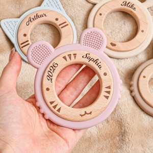 May include: A collection of baby teething toys in various animal shapes, including a cat, bear, and bunny. Each toy features a wooden ring with a silicone outer ring in pastel colors. The toys are personalized with names and the year 2026.