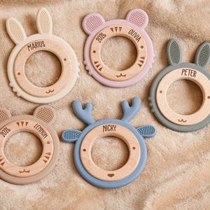 May include: Five baby teethers in various pastel colors, each with a wooden center and a unique animal shape: bunny, bear, and deer. Each teether has a name engraved on it, such as "Marius" and "Peter".