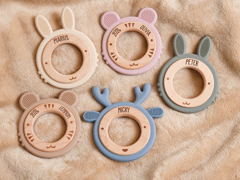 May include: Five baby teethers in various pastel colors, each with a wooden center and a unique animal shape: bunny, bear, and deer. Each teether has a name engraved on it, such as "Marius" and "Peter".